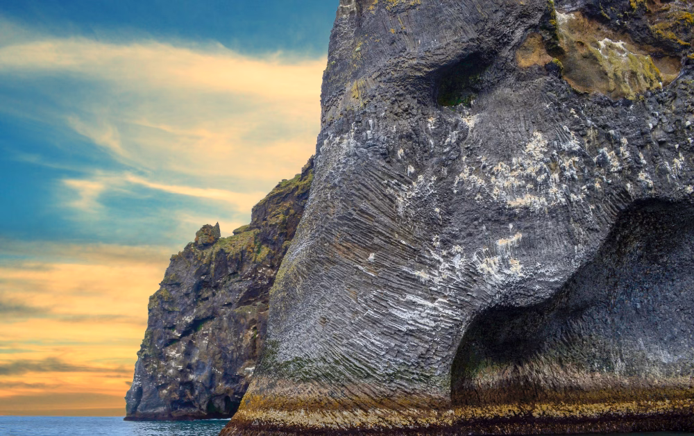 Basalt sea cliff shaped like an elephant’s head with a trunk dipping into the ocean at sunset.